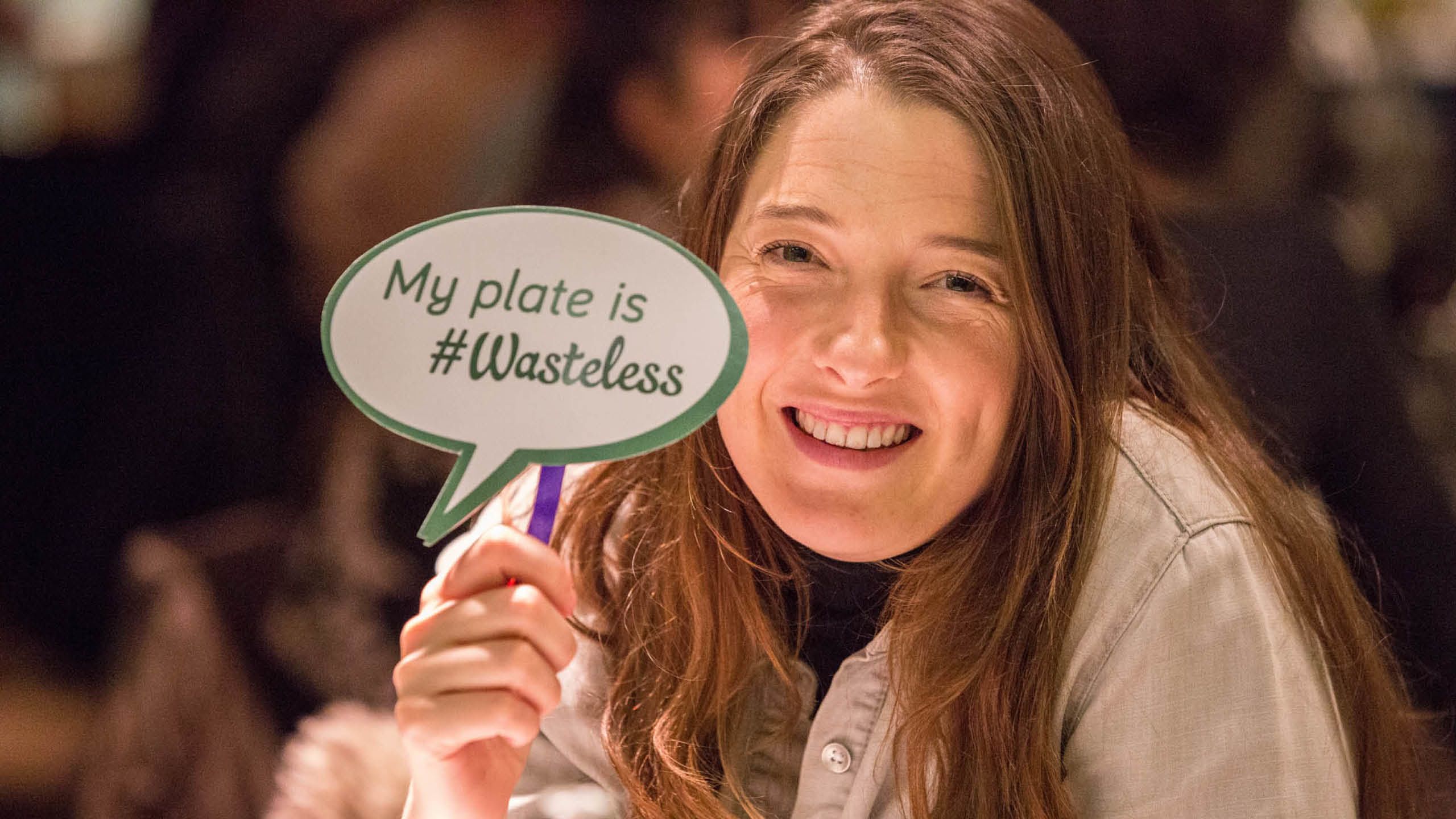 A diner holds up a paper speech bubble reading: My plate is #Wasteless
